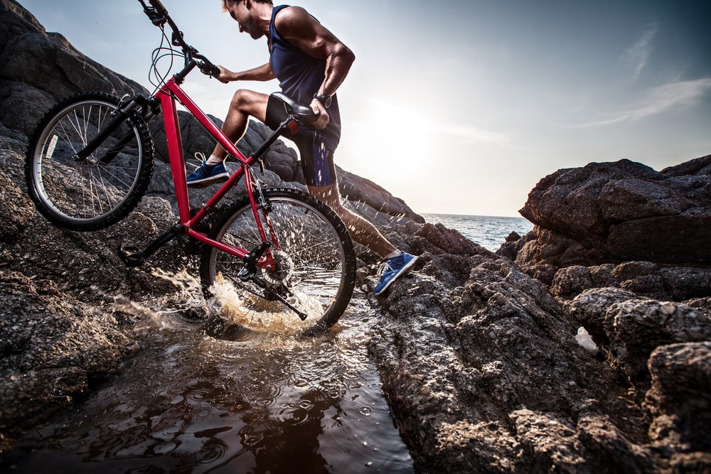 Man carrying a bicycle on rock across water during sunset