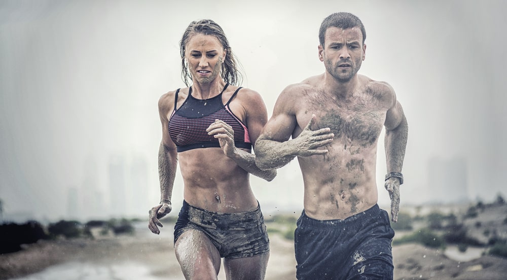 Muscular male and female athlete covered in mud running across rough terrain with a desert background